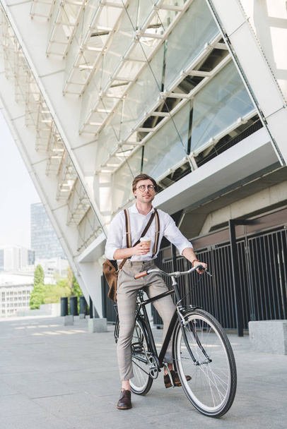 attractive young man with paper cup of coffee on vintage bicycle near sports stadium building - Photo, Image