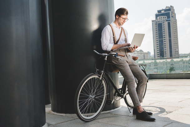 jovem elegante que trabalha com laptop enquanto se inclina na bicicleta vintage
 - Foto, Imagem
