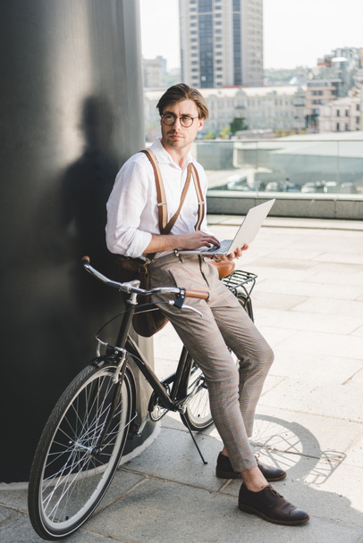 handsome young man working with laptop while leaning on vintage bicycle and looking away - Photo, image
