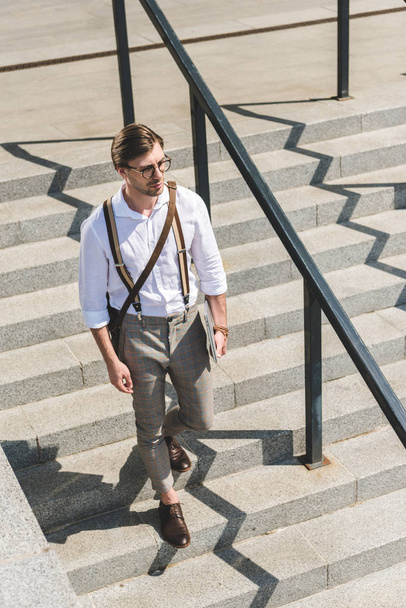 μεγάλη γωνία προβολής του όμορφος νεαρός άνδρας με το διπλωμένο laptop περπάτημα σε σκάλες σε δρόμο της πόλης - Φωτογραφία, εικόνα