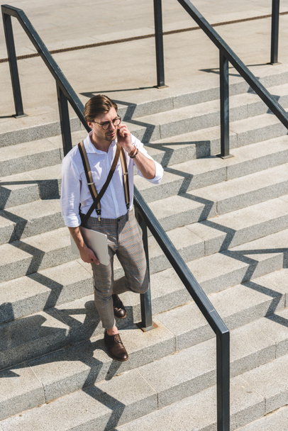 high angle view of stylish young man with folded laptop walking on stairs on city street and talking by phone - 写真・画像