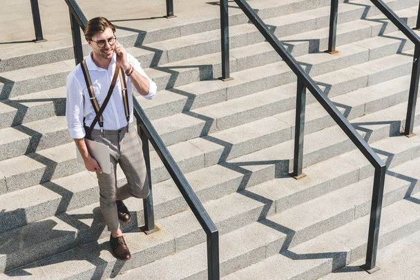 magas szög kilátás jóvágású fiatalember hajtogatott laptop járás lépcsőn a város utcája és a telefonon beszél - Fotó, kép