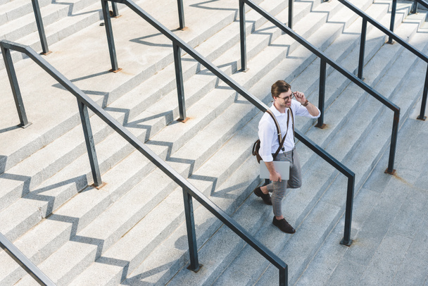 hoge hoekmening van stijlvolle jonge man lopen op trappen met laptop op stad straat - Foto, afbeelding