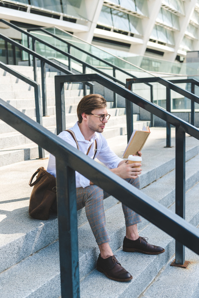 concentrated young man with paper cup of coffee reading book on stairs on city street - 写真・画像