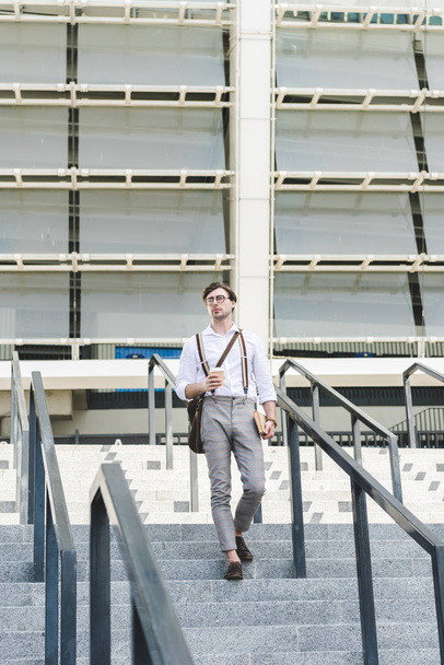 stylish young man walking down stairs in front of stadium with book and coffee to go - Zdjęcie, obraz