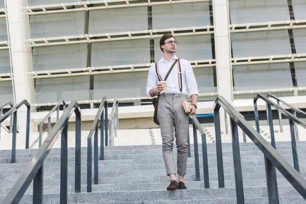 attractive young man walking down stairs in front of stadium with book and coffee to go - Photo, Image