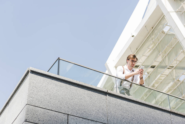 vista inferior de joven atractivo usando teléfono inteligente en balcón
 - Foto, Imagen