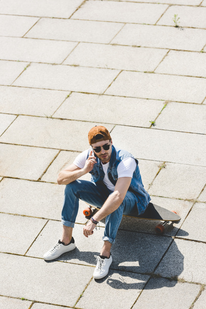 high angle view of handsome young skater sitting on longboard and talking by phone - Foto, immagini