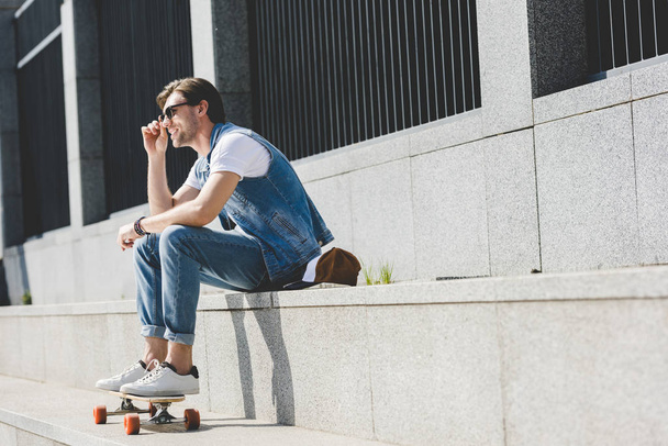 aantrekkelijke jonge man zittend op trap met skateboard weg op zoek - Foto, afbeelding