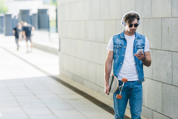 handsome young man in headphones with skateboard and smartphone - Photo, Image