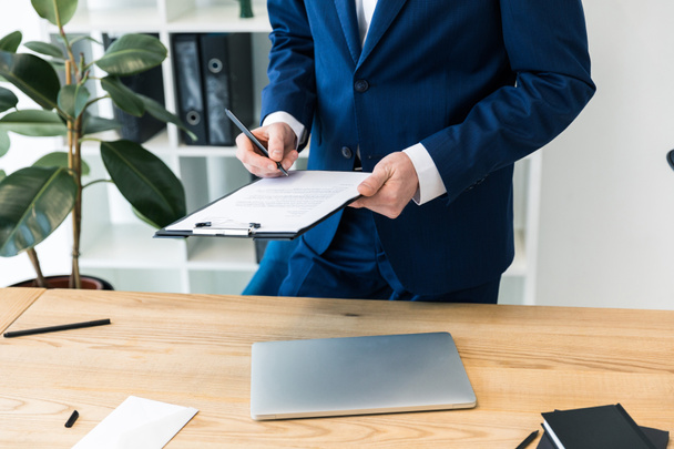 обрезанный снимок бизнесмена с блокнотом и ручкой в руках на рабочем месте с ноутбуком в офисе
 - Фото, изображение