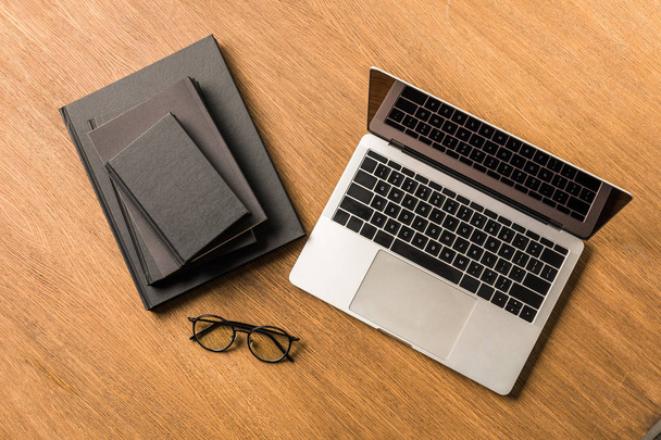 Draufsicht auf einen Stapel schwarzer Notizbücher, Laptops und Brillen auf einer hölzernen Tischplatte - Foto, Bild