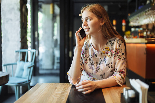 Young charming girl talking on the phone sitting alone in the cafe in her spare time, attractive woman with a cute smile having a conversation with a mobile phone while relaxing in the cafe - Foto, Bild