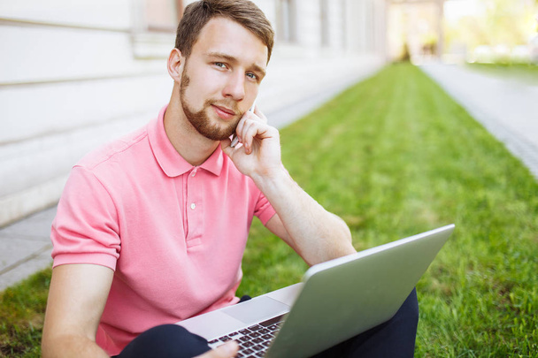 Handsome man sitting on the grass in the city with a laptop and talking on the phone, job search - Fotó, kép