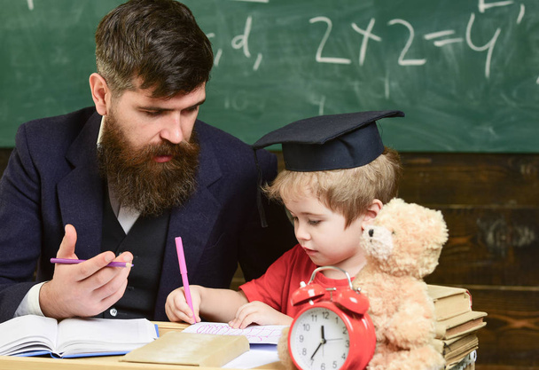 Elementary education. Teacher in formal wear and pupil in mortarboard in classroom, chalkboard on background. Kid studying with teacher. Father teaches son, discuss, explain. Education concept - Zdjęcie, obraz