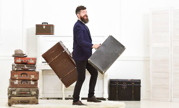 Man with beard and mustache wearing classic suit delivers luggage, luxury white interior background. Macho attractive, elegant on smiling face carries vintage suitcases. Butler and service concept - Foto, afbeelding