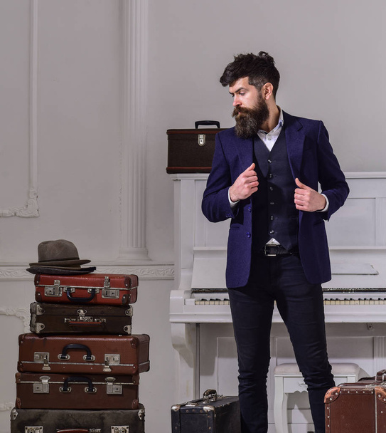 Macho stylish on thoughtful face standing near pile of vintage suitcase. Man, traveller with beard and mustache with luggage, luxury white interior background. Baggage and travelling concept - Foto, Imagen