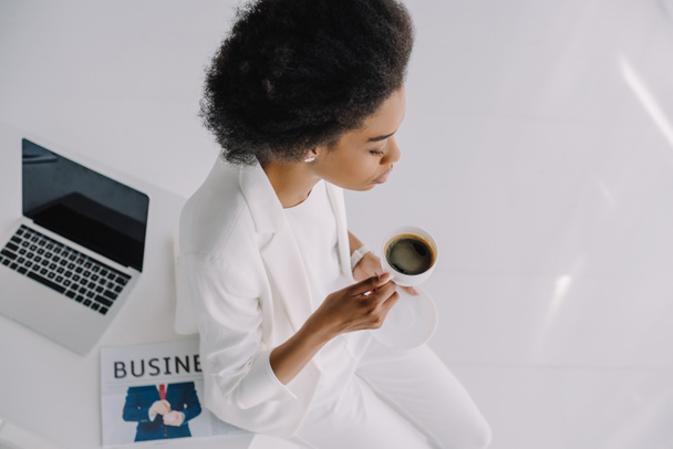 high angle view of attractive african american businesswoman sitting on table with cup of coffee in office - Fotó, kép