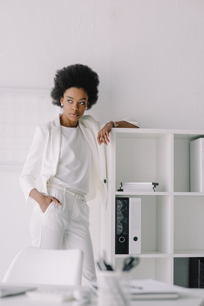 beautiful african american businesswoman leaning on shelves and looking away in office - Photo, Image