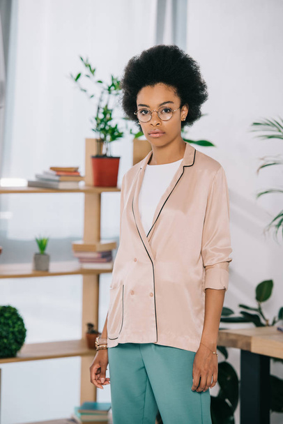 attractive african american businesswoman standing near table in office and looking at camera - Foto, afbeelding