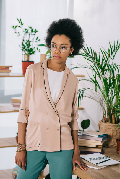 atractiva mujer de negocios afroamericana de pie cerca de la mesa en la oficina y mirando hacia otro lado
 - Foto, imagen