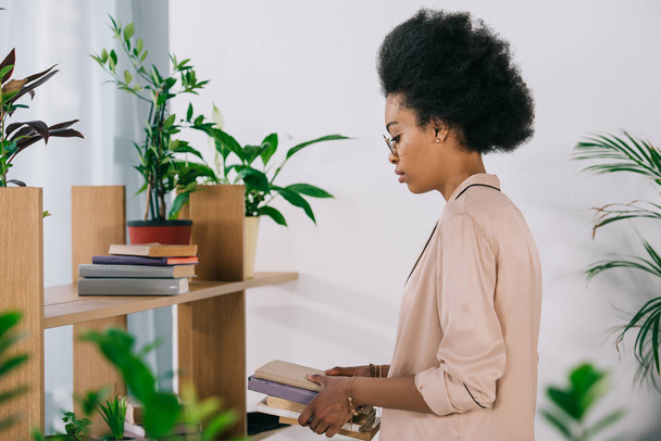 side view of attractive african american businesswoman holding books in office - Foto, Imagem
