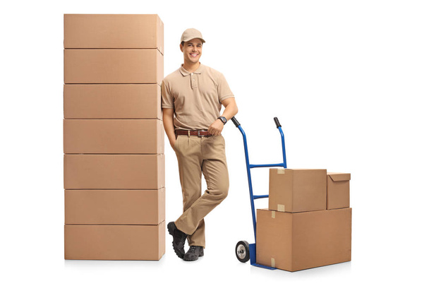 Full length portrait of a delivery man with a hand truck leaning on a pile of boxes isolated on white background - Фото, зображення