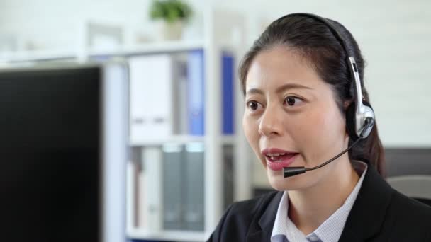 glücklich lächelndes asiatisches Mädchen im Anzug, das mit dem Kopfhörer im Büro am Computer sitzt. Call Center - Filmmaterial, Video