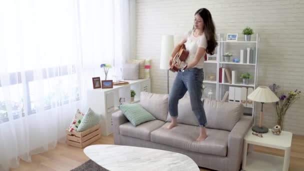 The quick movement of an Asian woman standing on the sofa playing guitar and dancing. - Imágenes, Vídeo