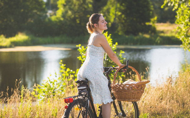 Τονισμένο πορτρέτο του όμορφη νεαρή γυναίκα στο σύντομο καλοκαιρινό φόρεμα ιππασία ποδήλατο στο ποτάμι - Φωτογραφία, εικόνα