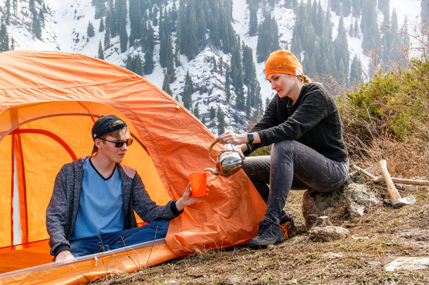 girl and a guy at a stop with a tourist tent drinking tea or coffee on a background of forest snow-capped mountains. A walking metal kettle. Leisure. Tourism. Healthy lifestyle - Photo, Image
