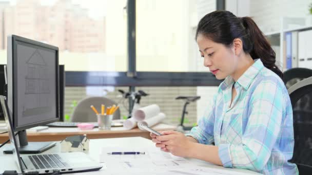 Képzett női építész beszélgető-val ügyfél keresése részletesen munka feladat mellett használ smartphone, miközben ő férfi munkatárs dolgozik a háttérben. - Felvétel, videó