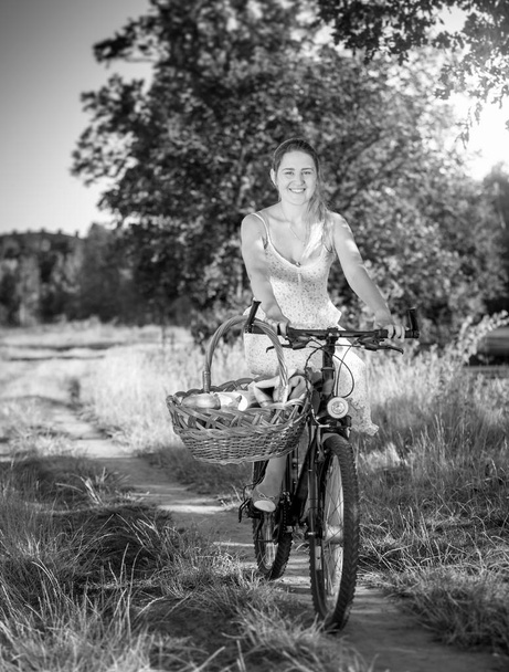 Black and white image of beautiful smiling woman riding on bicycle with big basket for picnic in park - Photo, Image