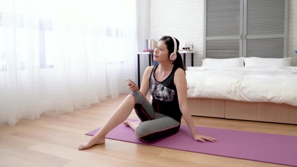 Asian woman sitting on the yoga mat and using her cell phone listening to the music. - Πλάνα, βίντεο
