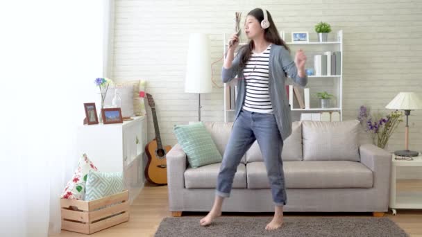 Asian woman sings with her cellphone as the microphone and danced in the living room. She follows the sounds in the headset and signs out the lyrics loudly. - Video