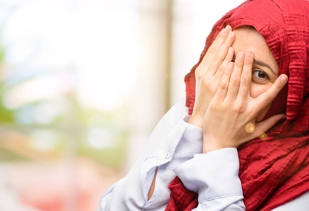 Young arab woman wearing hijab smiling having shy look peeking through her fingers, covering face with hands looking confusedly broadly - Фото, изображение