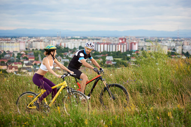 Δύο ποδηλάτες χαρούμενος - άνδρας και η γυναίκα ιππασία σε ένα λόφο στο το γρασίδι με άγρια λουλούδια, πόλη και τα βουνά στο βάθος. - Φωτογραφία, εικόνα
