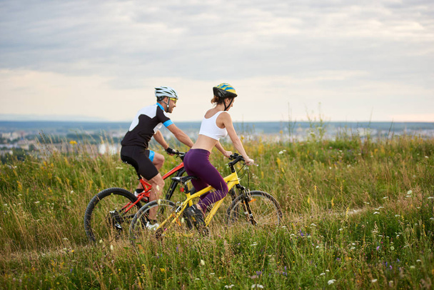 Δύο ποδηλάτες χαρούμενος - άνδρας και η γυναίκα ιππασία σε ένα λόφο στο το γρασίδι με άγρια λουλούδια, πόλη και τα βουνά στο βάθος. - Φωτογραφία, εικόνα