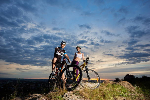 Χαμογελώντας νεαρό ζευγάρι των ποδηλατών σε κράνη κάθεται σε ποδήλατα βουνού και κοιτάζοντας κάμερα.  - Φωτογραφία, εικόνα