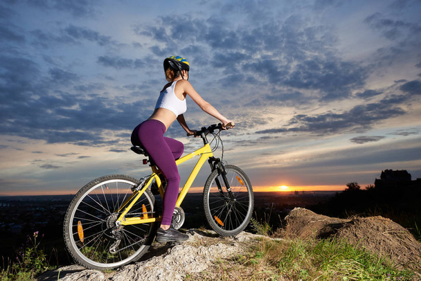 Sportowy kobieta z rowerów, pozowanie na wzgórzu. Szczupła i piękna kobieta nosi fioletowe leginsy i biały top.  - Zdjęcie, obraz