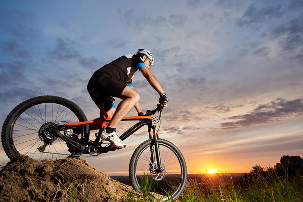 Active male cyclist wearing sportswear and helmet, riding bicycle alone and rolling down hill. Sporty and robust man cycling against beautiful sunset and rose-blue sky in the evening background - 写真・画像