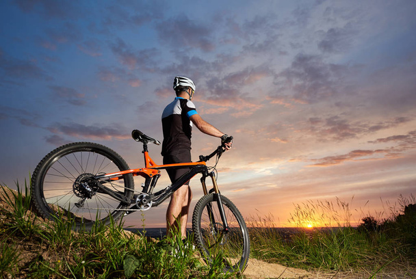 Vista posterior de ciclista masculino en un casco y ropa deportiva descansa sobre el fondo de una puesta de sol desde una altura observando la montaña en la distancia, el cielo encantador y el sol brillante
 - Foto, imagen
