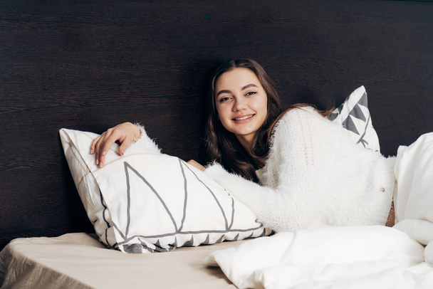 beautiful long-haired girl in white pajamas lies in bed early in the morning, smiling, good day - Фото, изображение