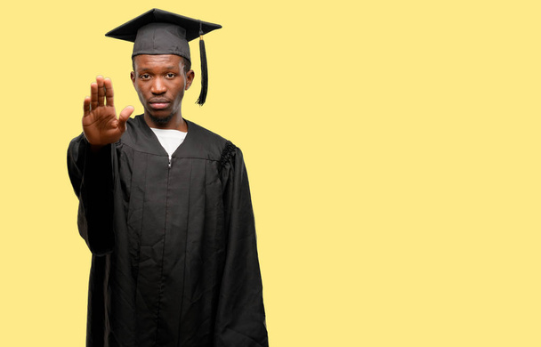 Giovane africano studente laureato uomo di colore infastidito con cattivo atteggiamento facendo segno di stop con la mano, dicendo di no, esprimendo sicurezza, difesa o restrizione, forse spingendo
 - Foto, immagini