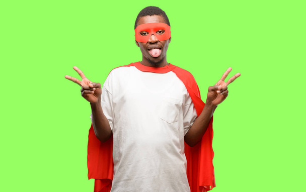 Afrikaanse zwarte superheld man kijken camera tonen van de tong en het teken van de overwinning met vingers - Foto, afbeelding