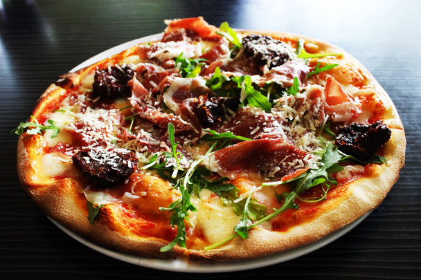 Pizzaa aurinkokuivatuilla tomaateilla, prosciuttolla, arugulalla ja parmesaanijuustolla pöydällä olevalla lautasella
 - Valokuva, kuva