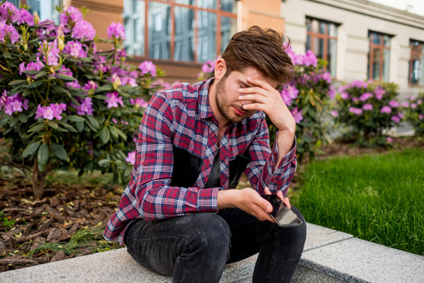 portrait of young sad bearded man in plaid shirt outdoor with pink flowers on background - Foto, Imagen