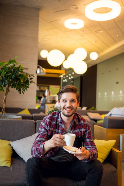 portrait of young bearded man in plaid shirt holding white cup of coffee indoor on grey sofa background  - Fotó, kép