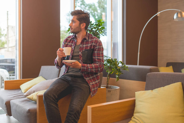 portrait of young bearded man in plaid shirt drink coffee from white cup indoor on grey sofa background - Photo, Image