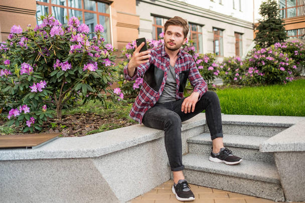 背景にピンクの花と屋外の携帯を使用して石の階段に座っている格子縞のシャツの若いひげを生やした男の肖像 - 写真・画像